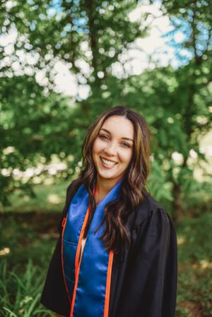 Graduate in academic dress smiles brightly during outdoor photo session with natural greenery.