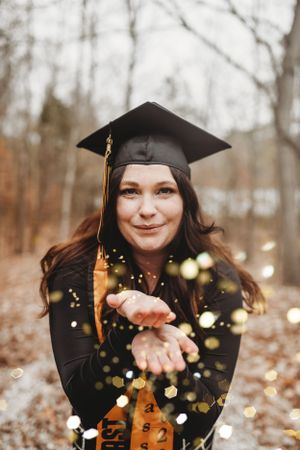 Graduate tossing gold glitter in celebration against winter forest backdrop.