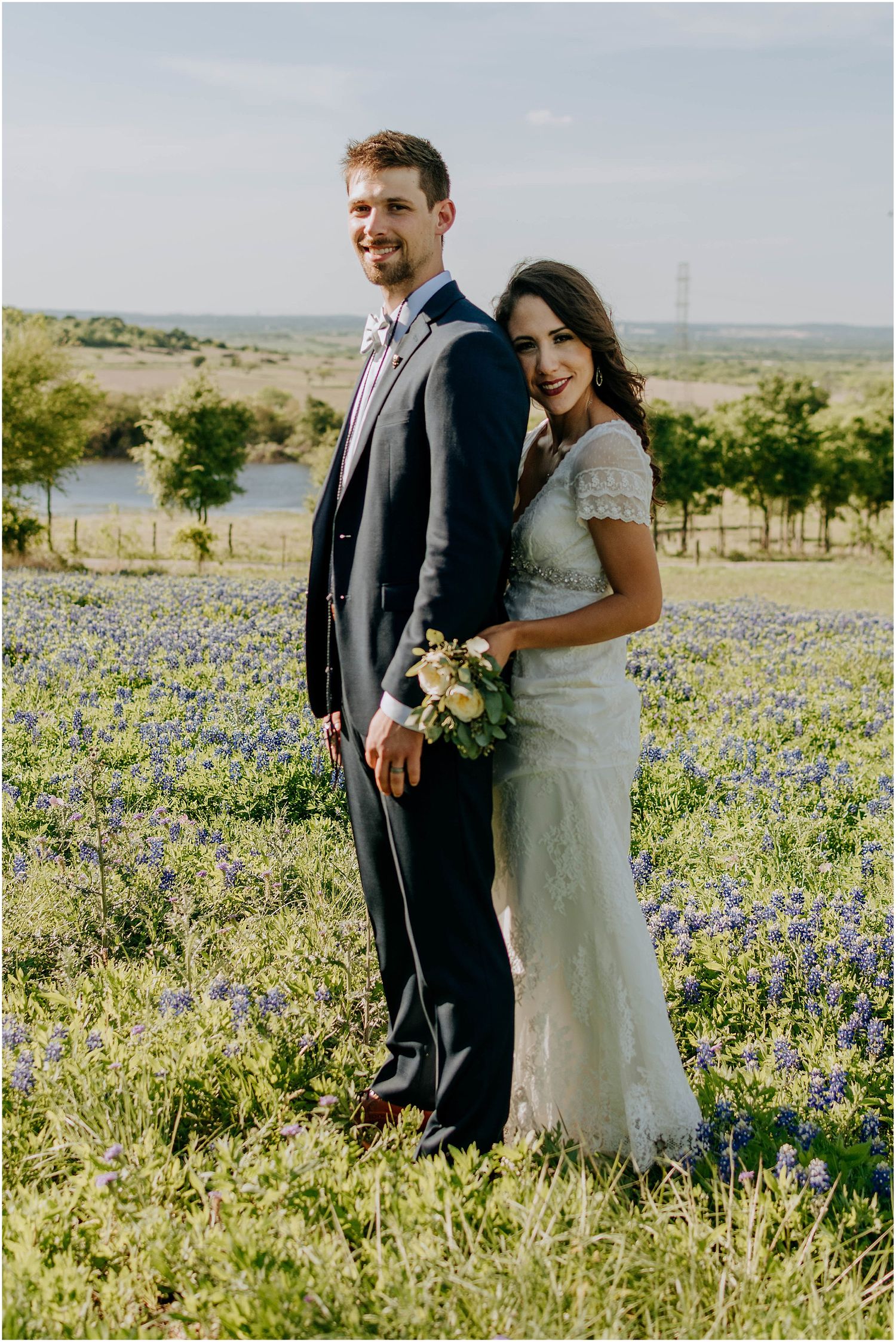 A newlywed couple poses in a scenic field of bluebonnets with a lake view behind them.