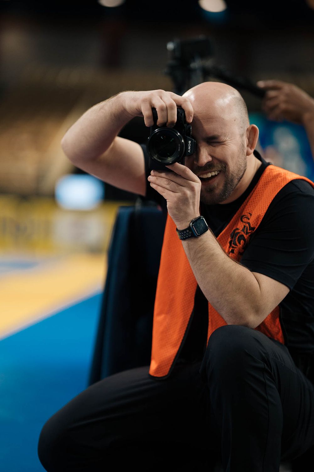 A photographer in an orange safety vest takes photos at an indoor sporting event.