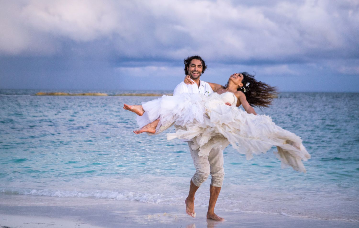 Couple celebrating their beach wedding with playful lift pose against turquoise waters.