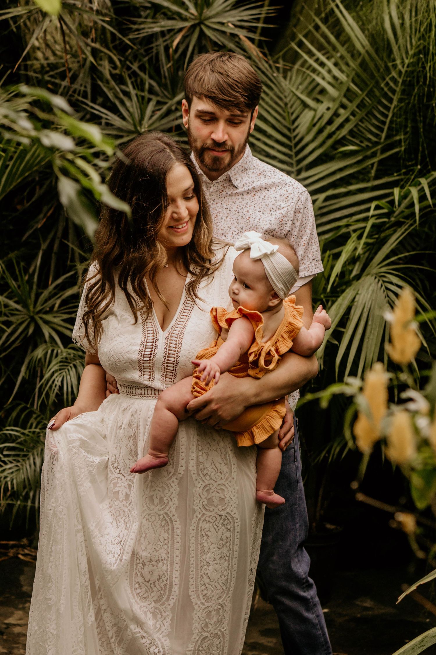 Young family with baby girl in orange dress posing among tropical plants in greenhouse setting.