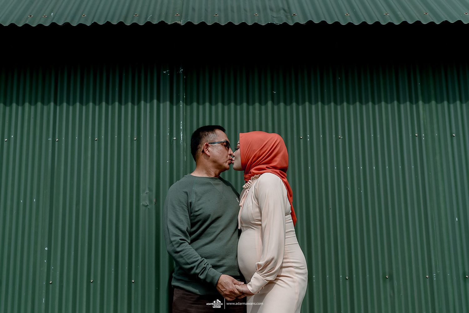 Couple posing against green corrugated metal wall in outdoor setting.