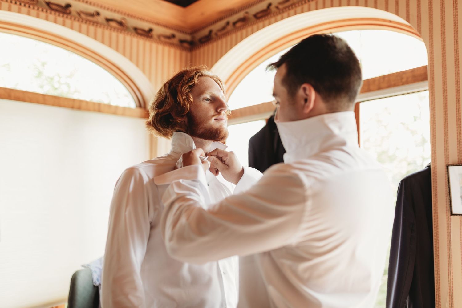 Two people in formal wear help adjust a bowtie near a bright window.