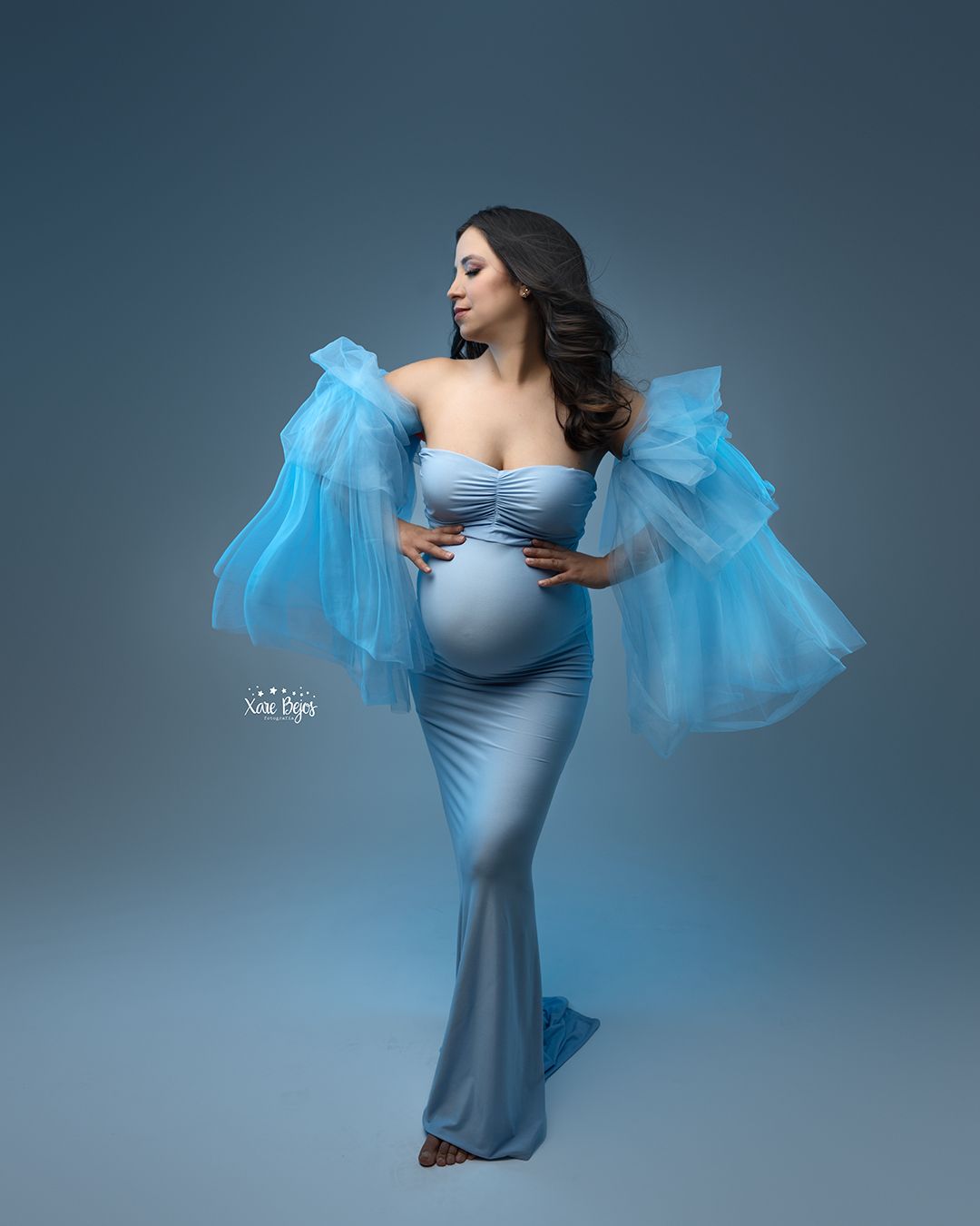 Maternity portrait with flowing blue tulle sleeves against dark backdrop.