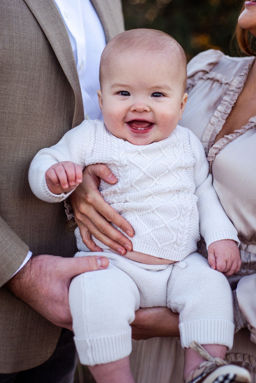 Baby wearing white knit sweater and pants smiling while being held in family photo session.