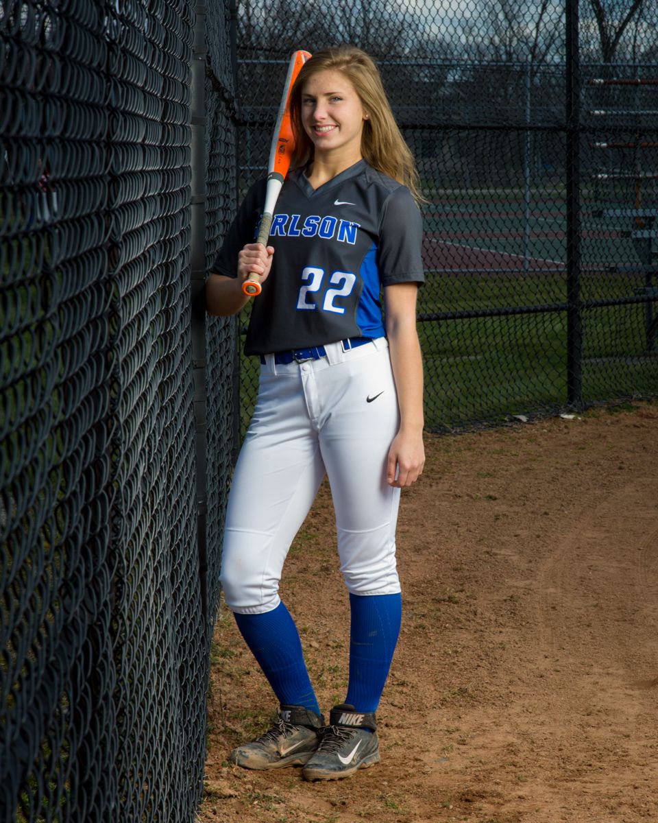 Female softball player in Carlson number 22 uniform leaning on a fence holding an orange bat.