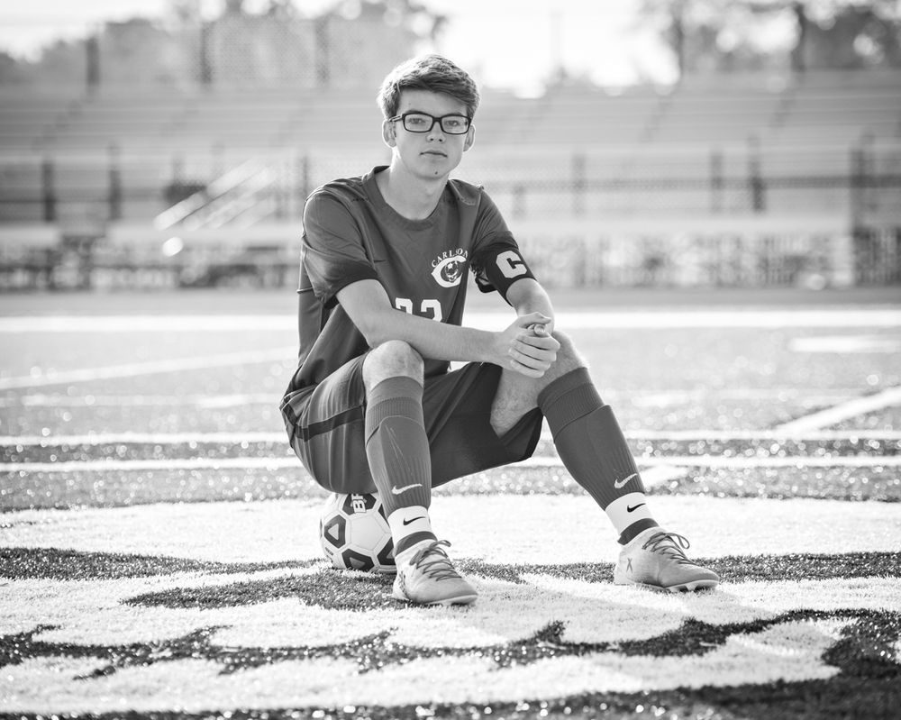 Black and white photo of young male soccer player sitting on stadium field turf.