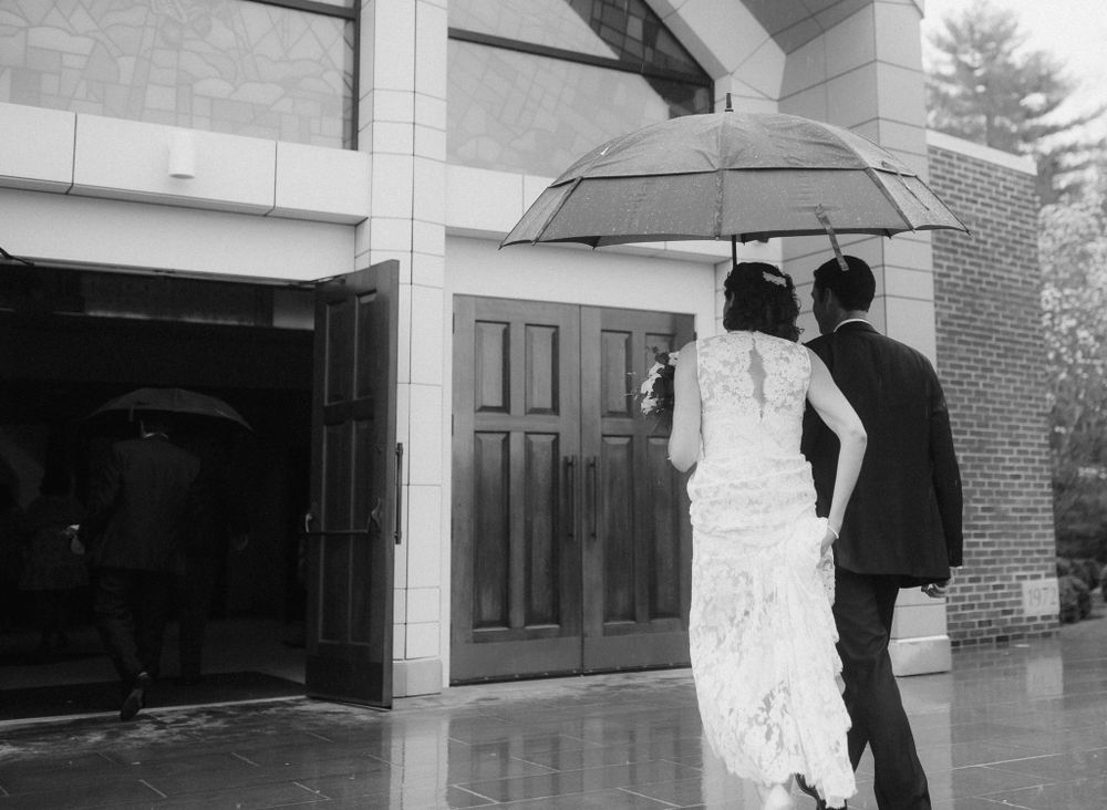 A couple under an umbrella walking towards church doors on a rainy day in black and white.