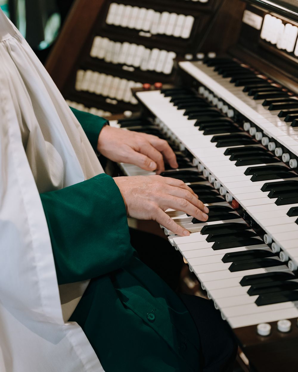Hands playing a church organ keyboard with white and black keys in green vestments.