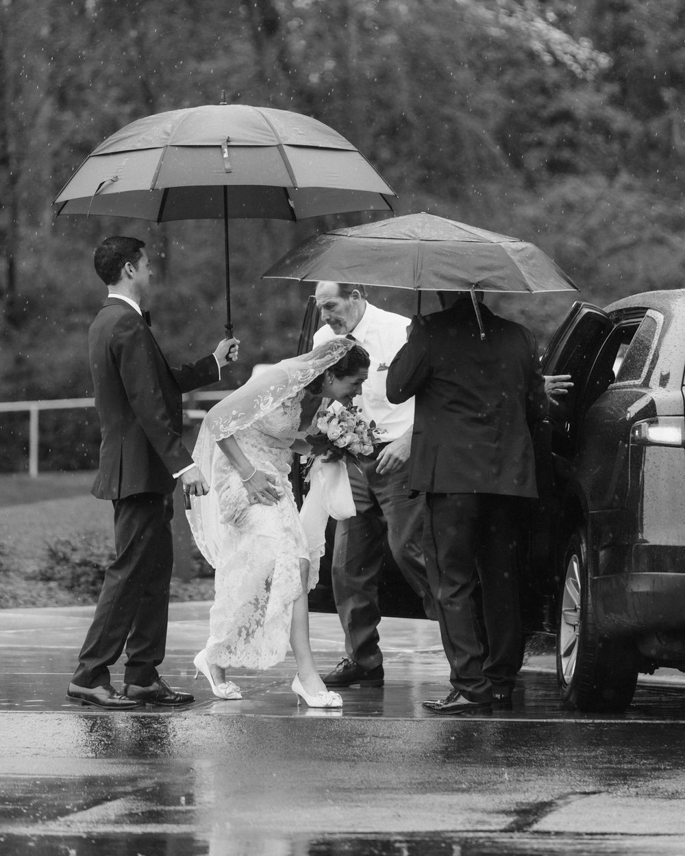 Wedding party huddles under black umbrellas in the rain during an outdoor ceremony.