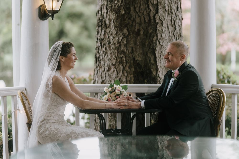 Newlyweds share intimate moment at outdoor table framed by rustic columns and trees.