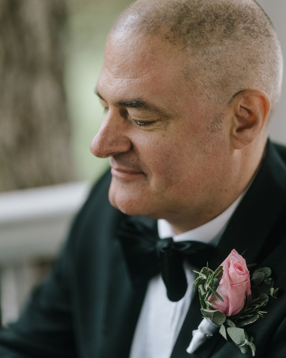 Groom in black tuxedo with pink rose boutonniere smiling in profile.