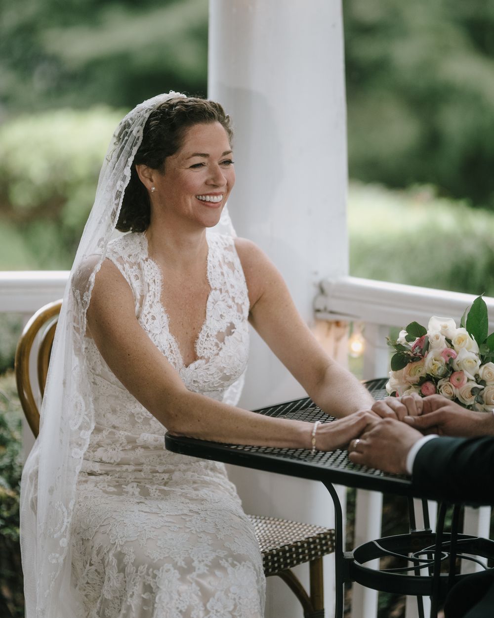 Bride in lace dress smiles while seated at outdoor table during wedding.