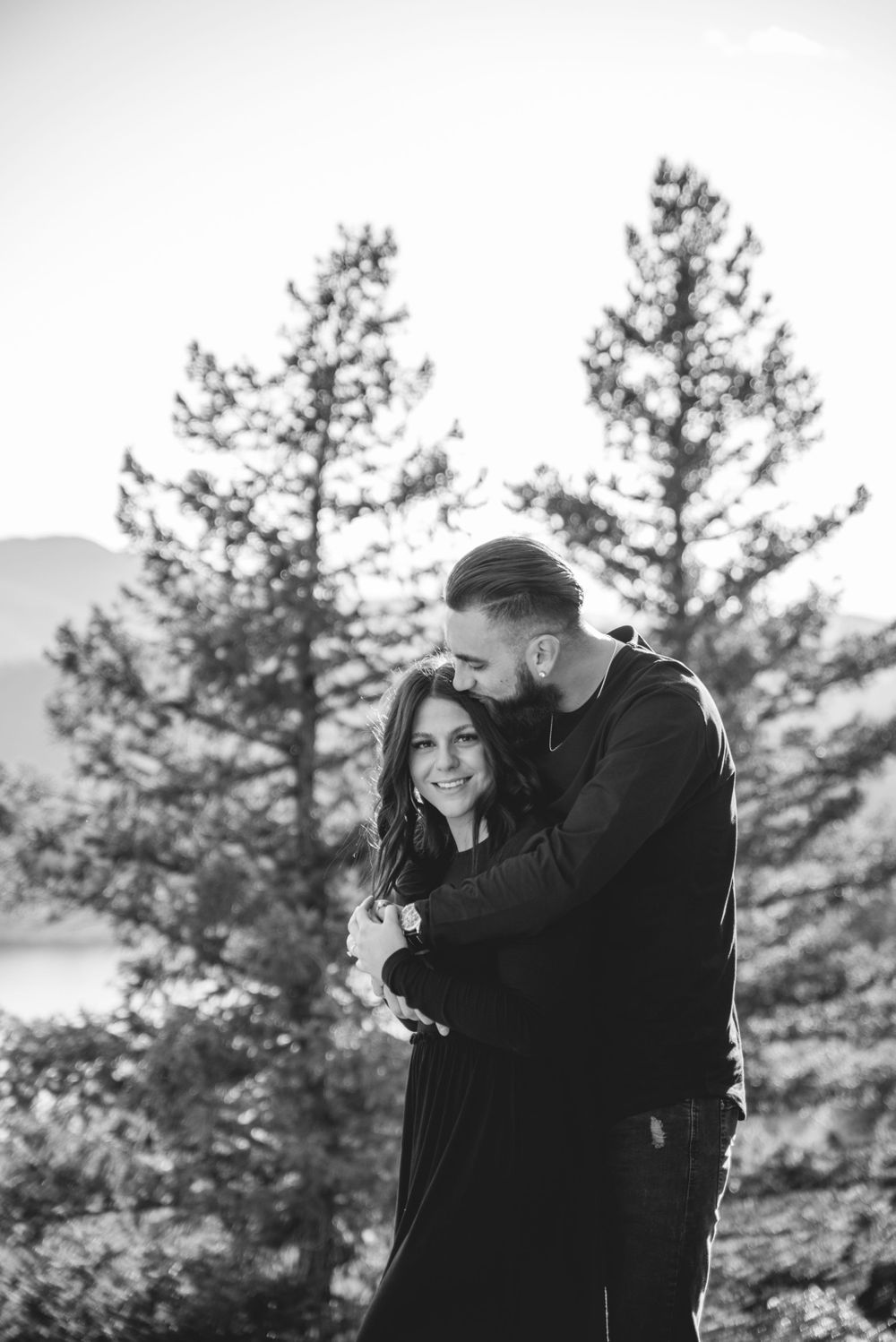A couple shares an intimate moment among pine trees with mountains in the background.