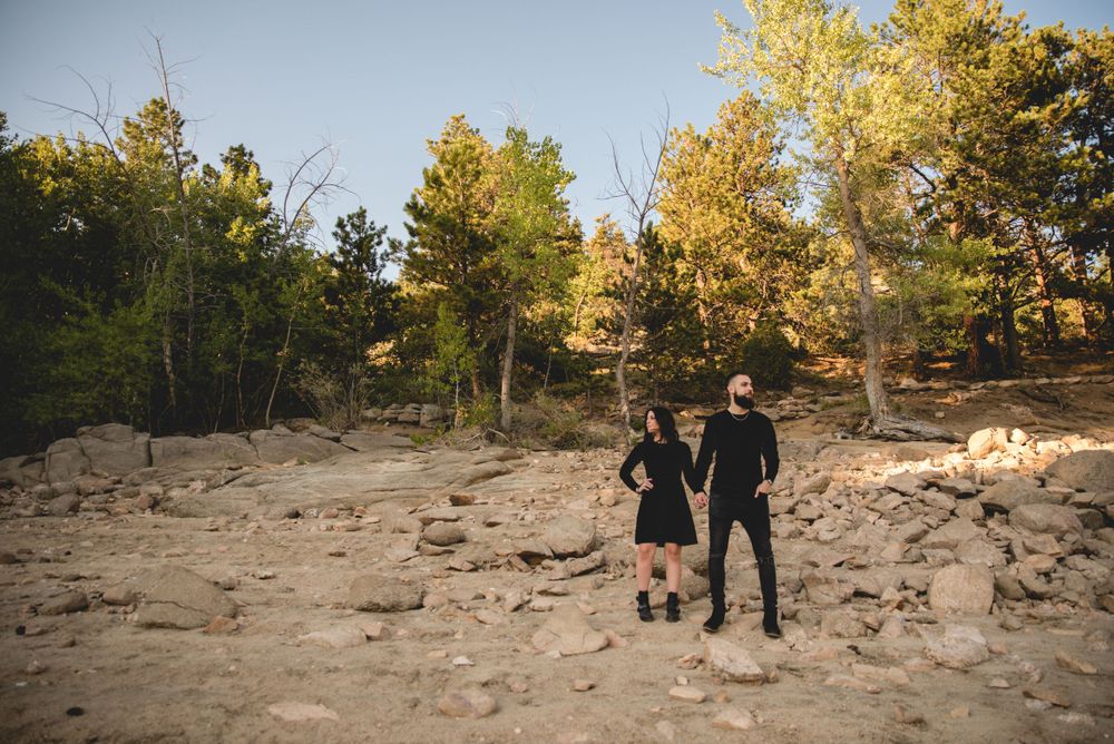 Couple walking hand in hand on rocky terrain with autumn trees in background.