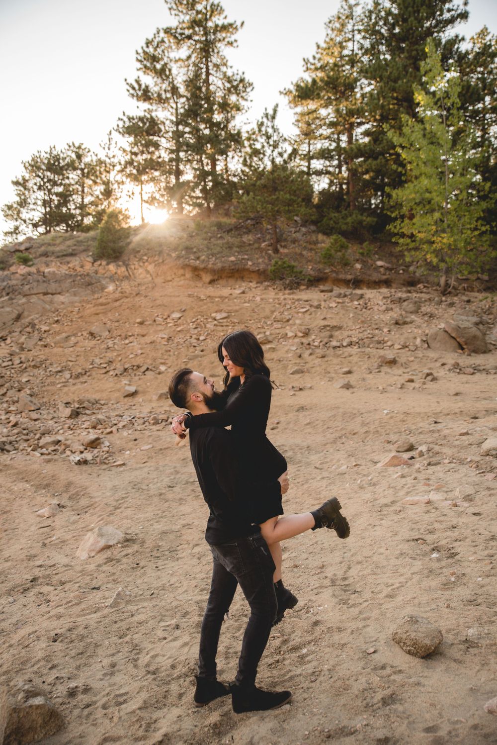 Couple sharing playful moment on hillside during sunset with pine trees in background.