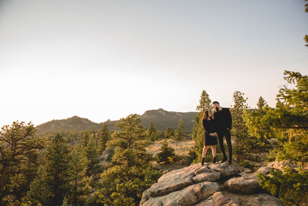 Couple standing on rocky outcrop overlooking mountain landscape at sunset.