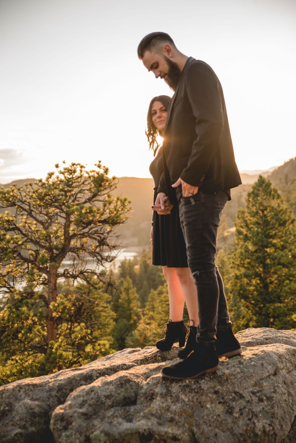 Couple standing together on rocky overlook with mountain scenery at sunset.