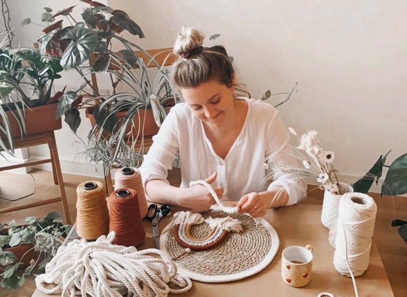 Woman crafting macrame at a wooden table surrounded by plants and colorful yarn spools in a cozy studio.