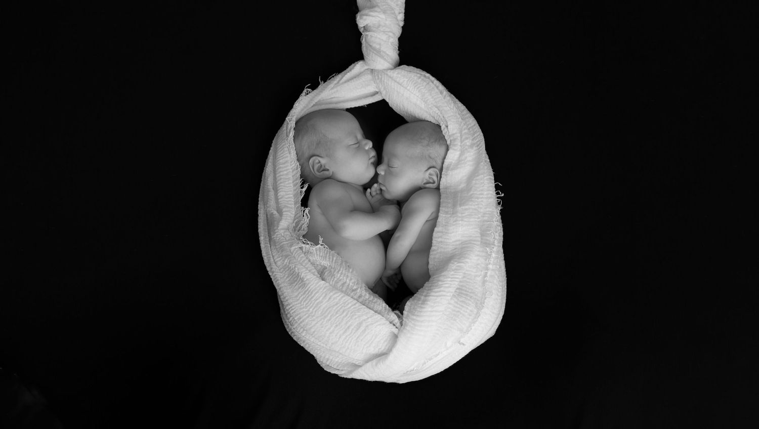 Black and white overhead view of newborn twins nestled together in soft fabric.