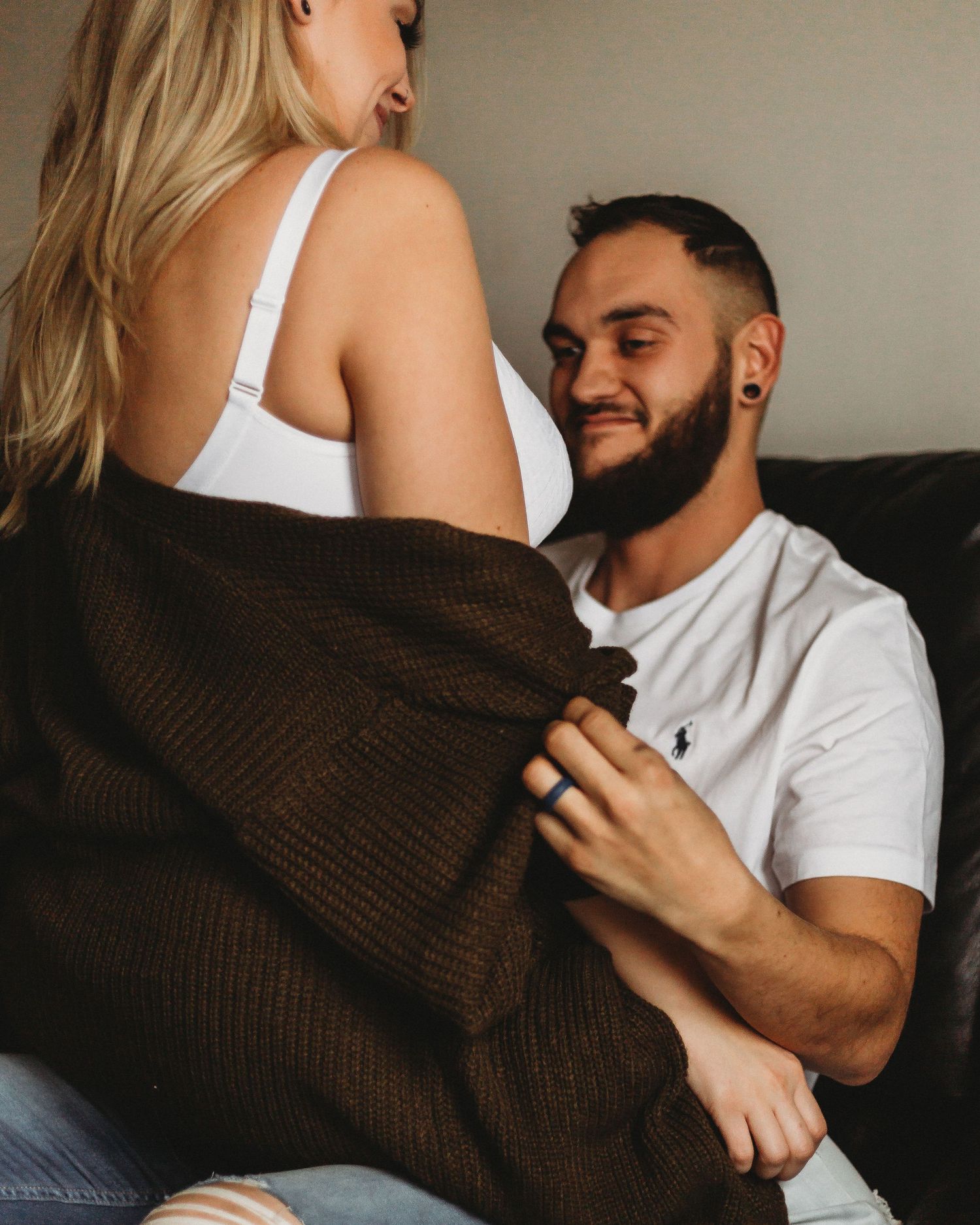 Couple sharing playful moment on couch wearing casual attire with brown sweater.