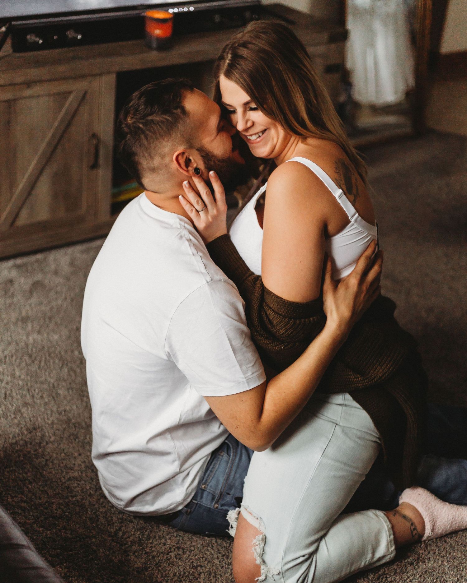 Playful couple sharing intimate moment on carpeted floor in casual white and brown outfits.