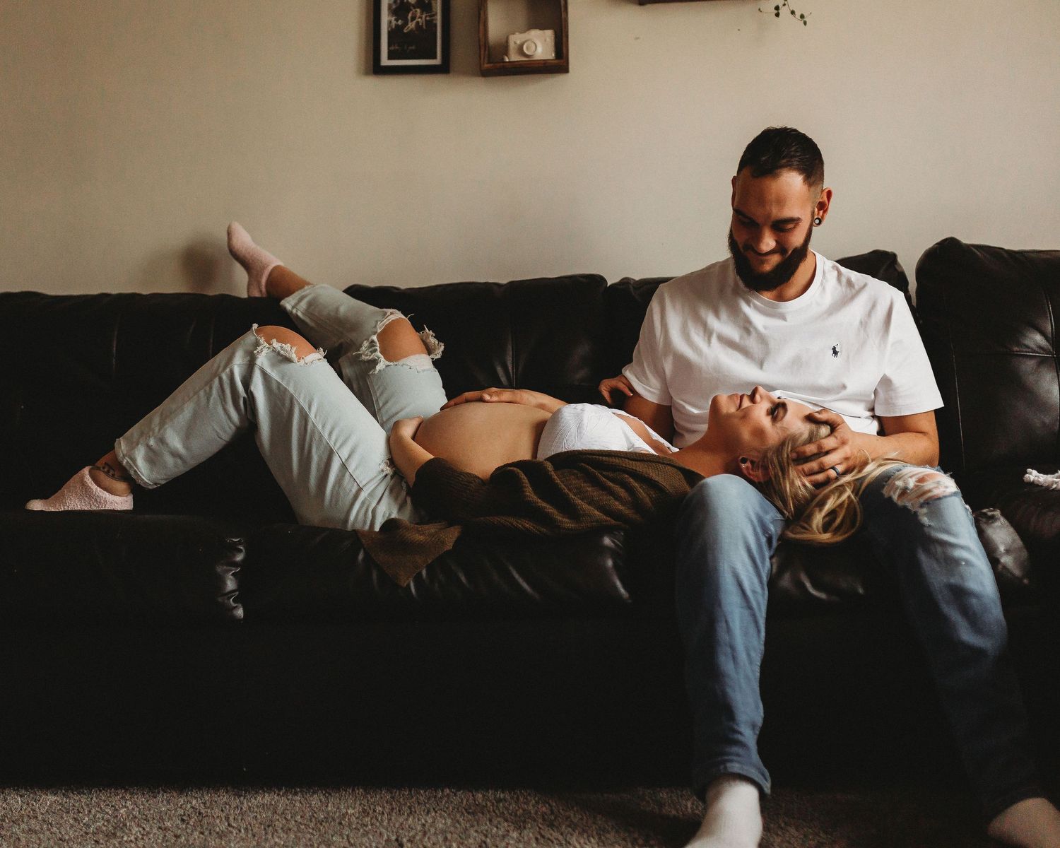Relaxed couple lounging on dark leather couch in casual white shirt and ripped jeans.