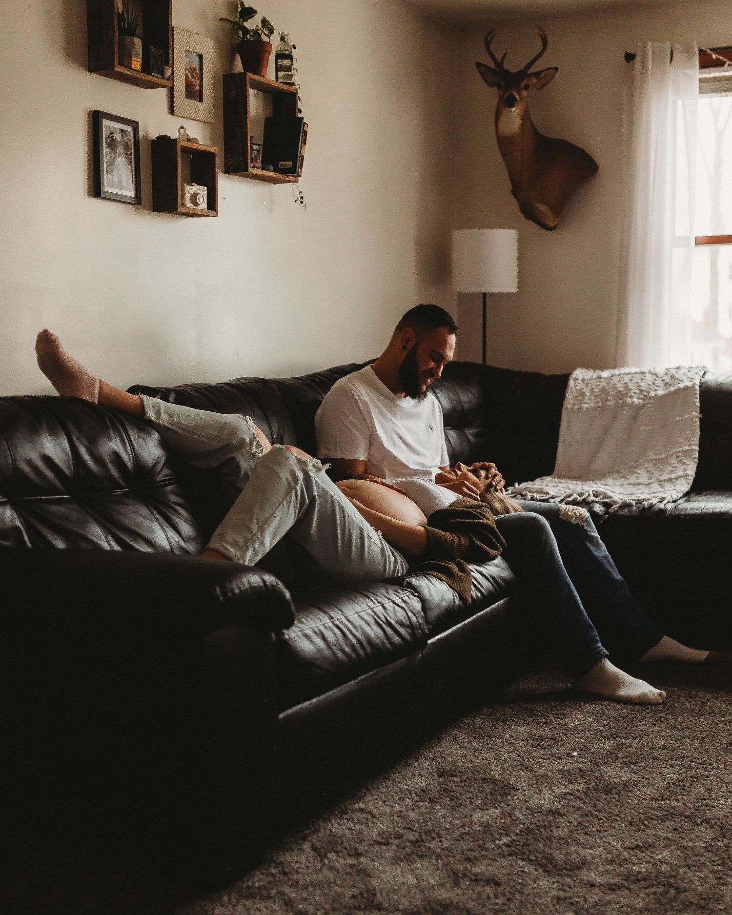 Relaxed couple sharing intimate moment on leather couch with mounted deer head and framed photos on wall.