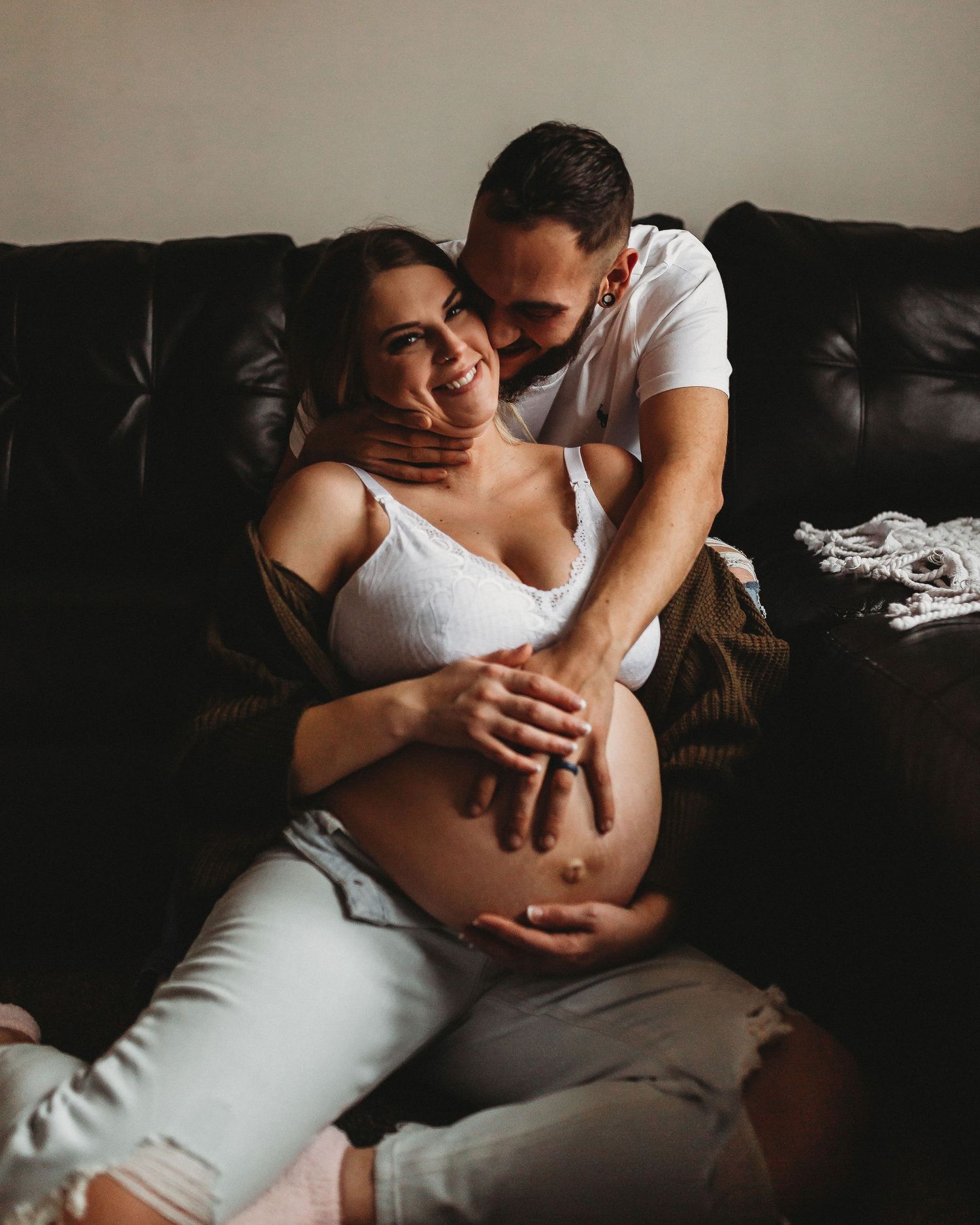 Intimate maternity photoshoot on leather couch showing couple embracing pregnant belly.
