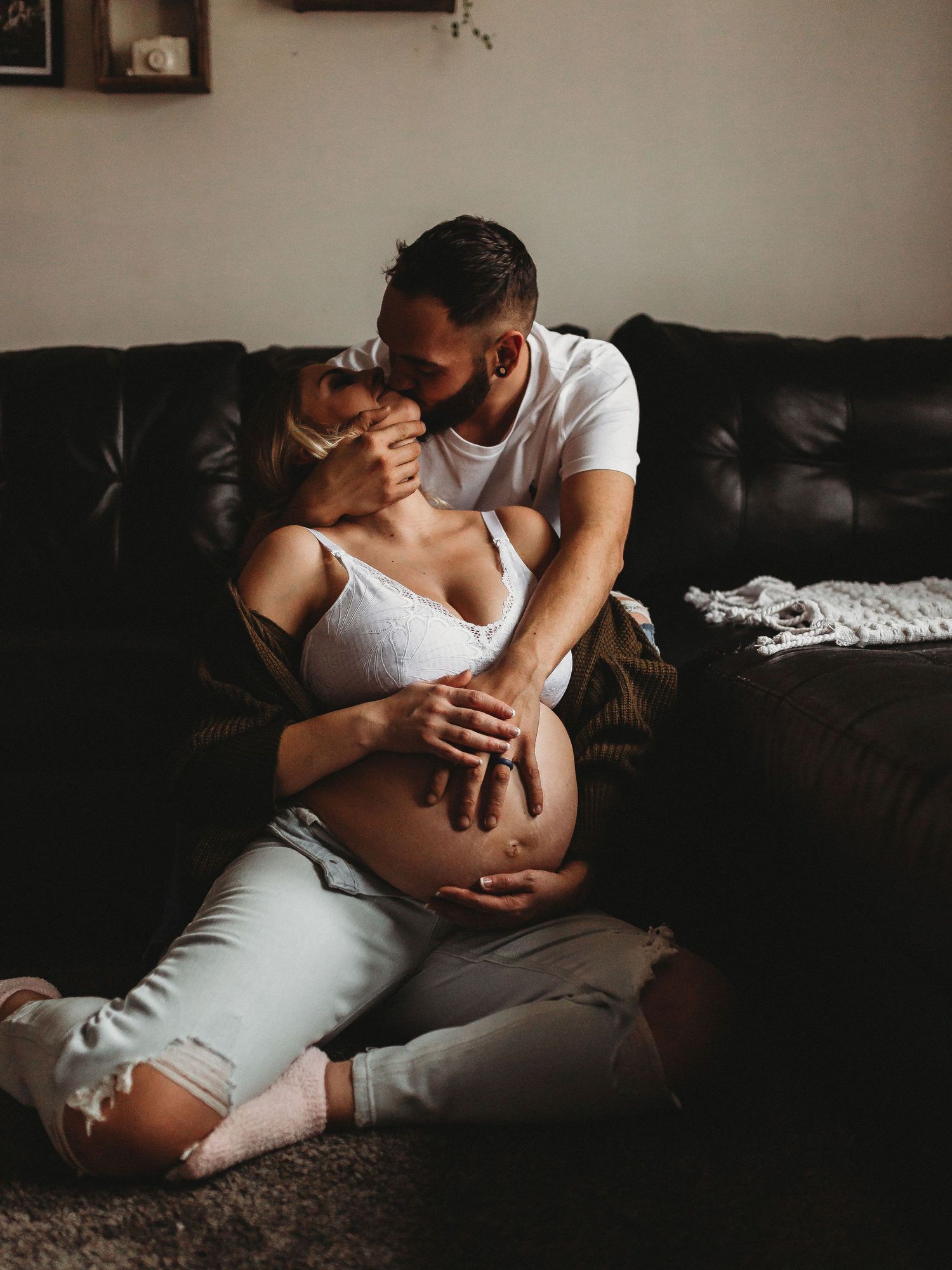 Romantic maternity moment captured on dark leather couch in natural lighting.