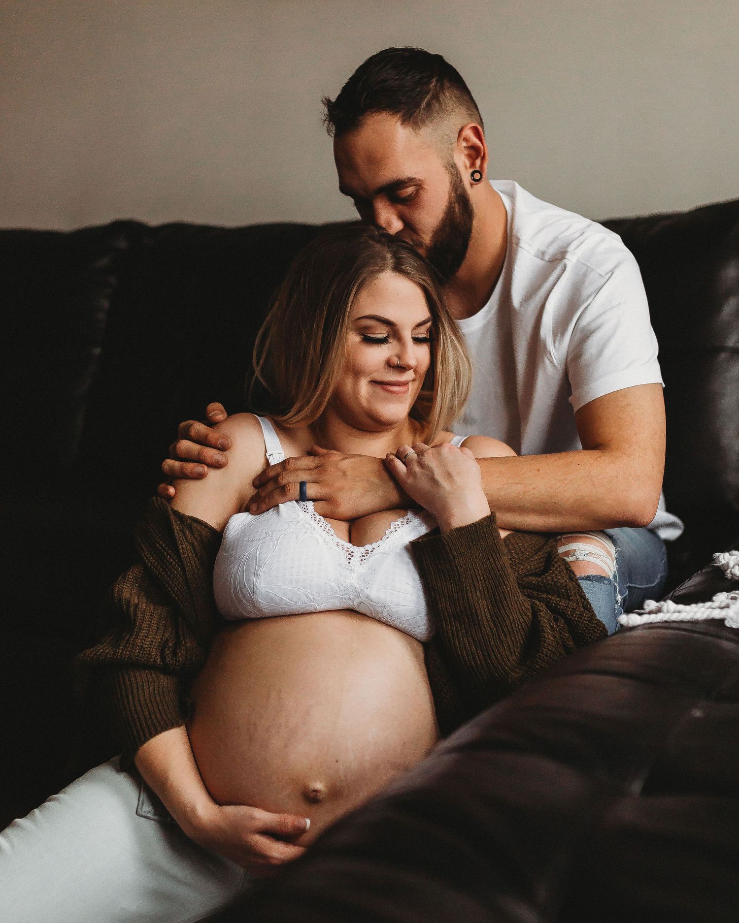 Sweet pregnancy photoshoot with couple cuddling on dark leather couch.