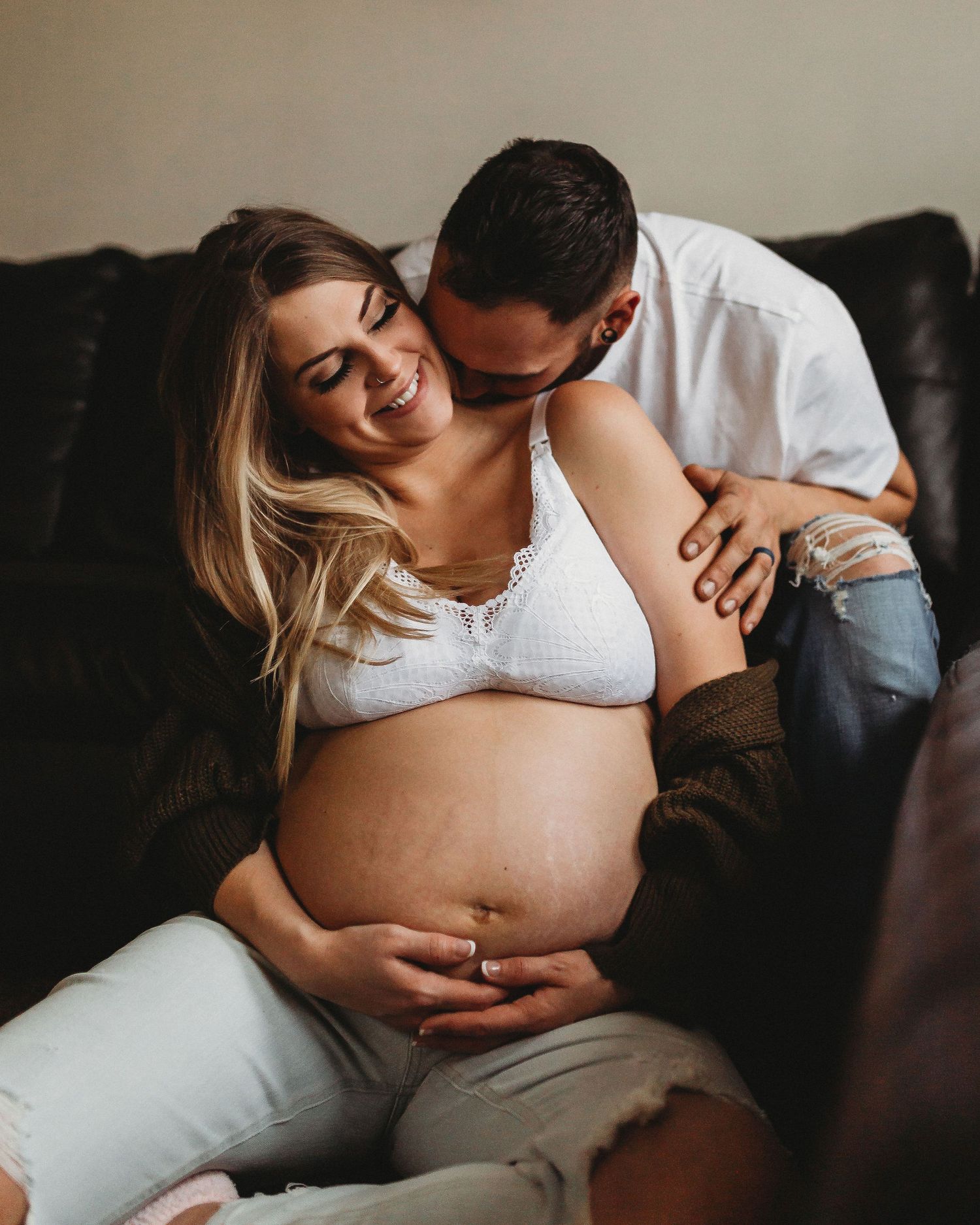 Joyful maternity moment captured with couple laughing on leather sofa.