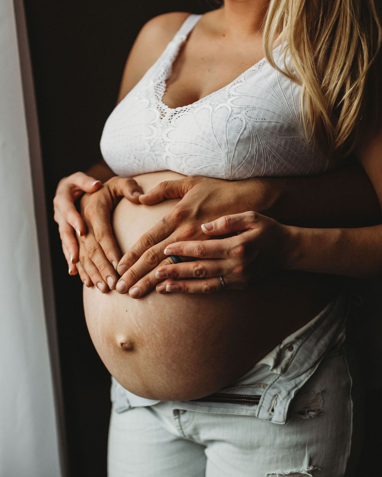 Side view of pregnant belly with hands placed lovingly against white lace bralette.