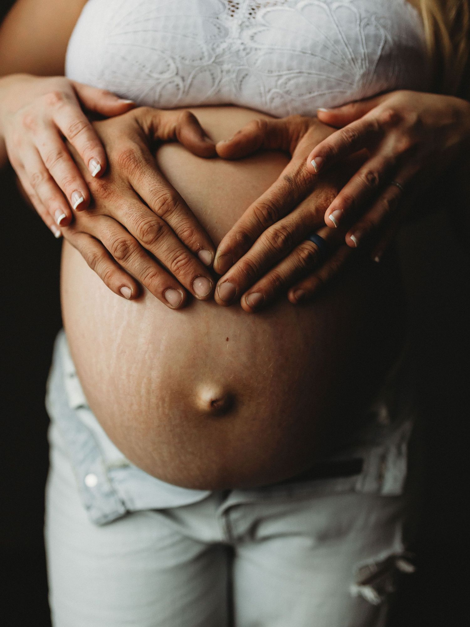 Close up of hands forming heart shape on pregnant belly in white lace top.