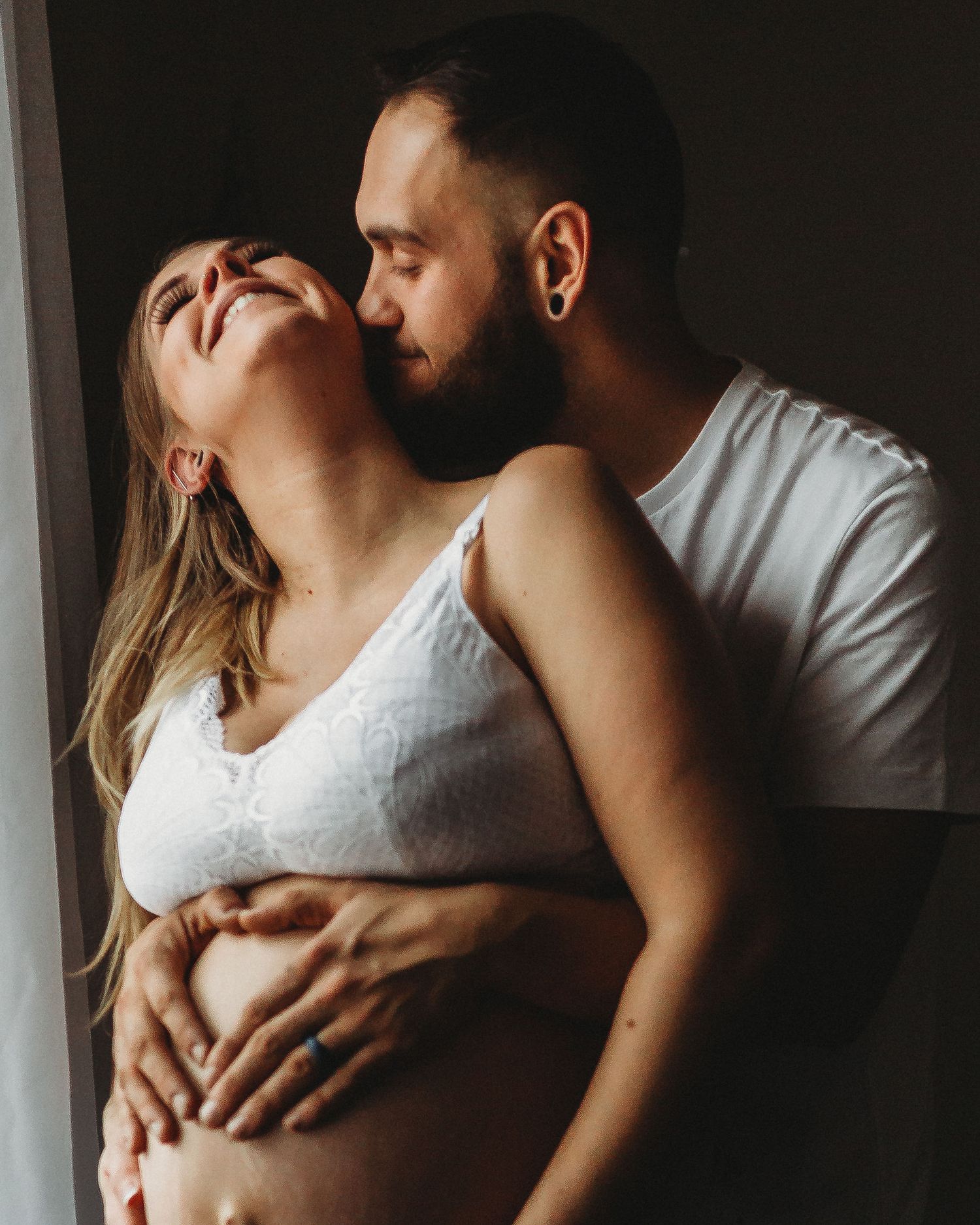 Intimate maternity portrait of couple sharing tender moment by window.