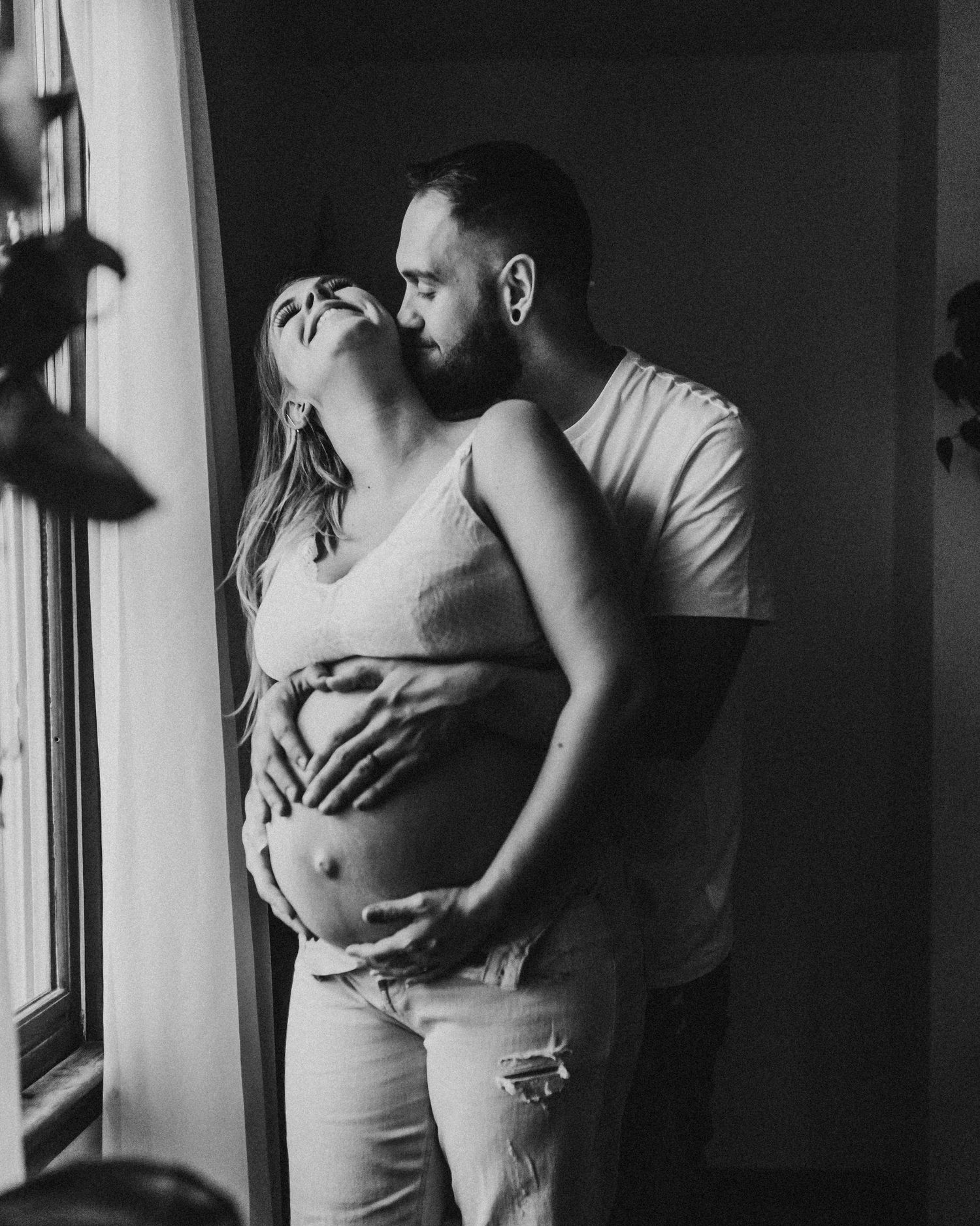 Intimate black and white maternity photo of couple embracing by window.