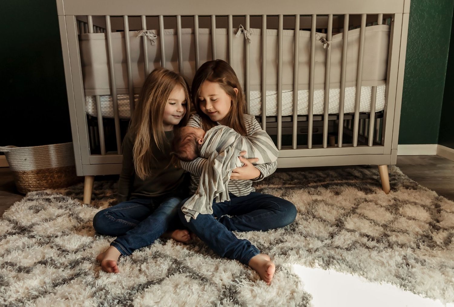 Siblings sitting on patterned rug in front of modern gray crib.