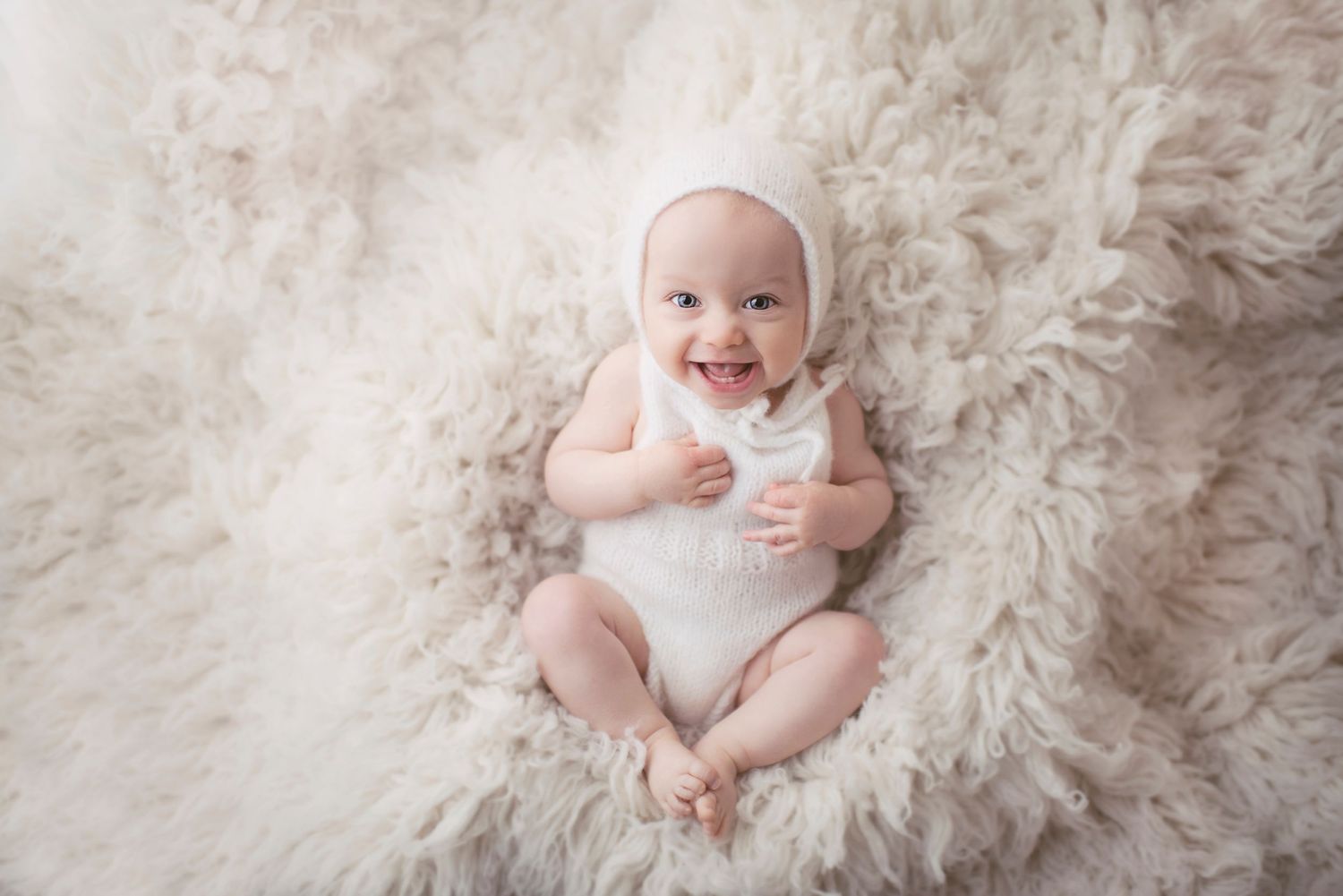 A joyful baby in white clothing lies on plush fur during a bright studio photoshoot.