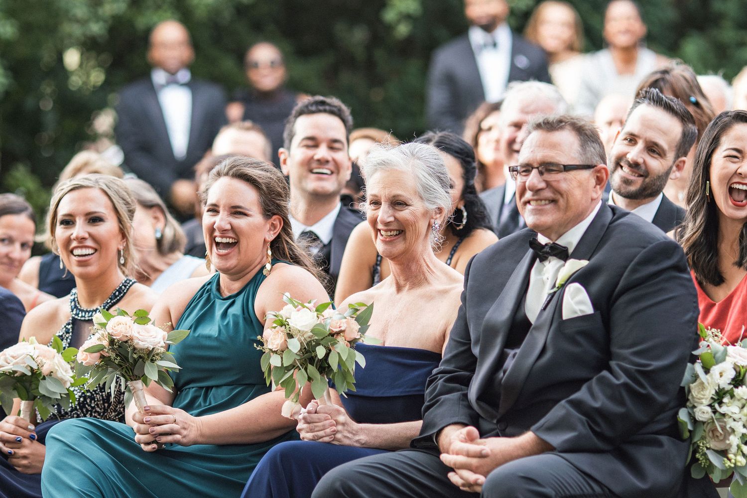 Wedding guests laughing together while seated in formal attire at an outdoor ceremony.