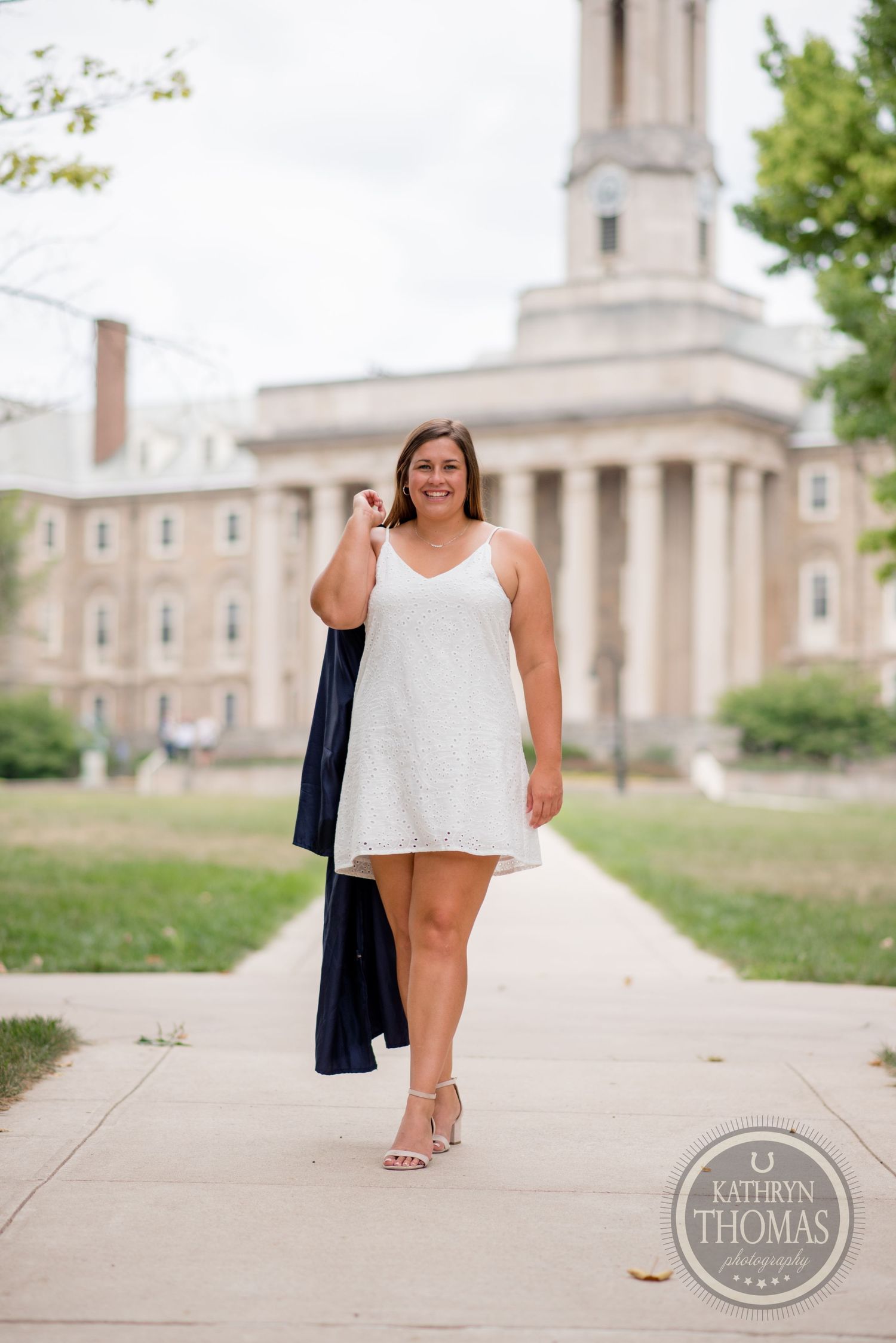 Graduate in white dress and academic regalia walks confidently past historic university building.