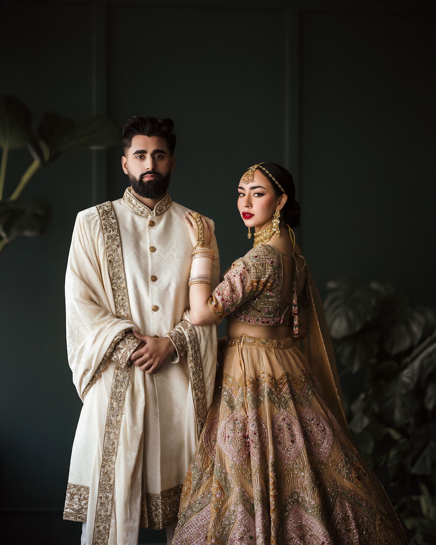 Couple in traditional South Asian wedding attire posing against dark backdrop.
