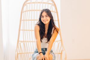 Person posing cheerfully in a woven rattan chair against pink wall.