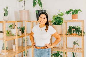 Woman standing next to wooden shelving unit filled with houseplants in bright interior.