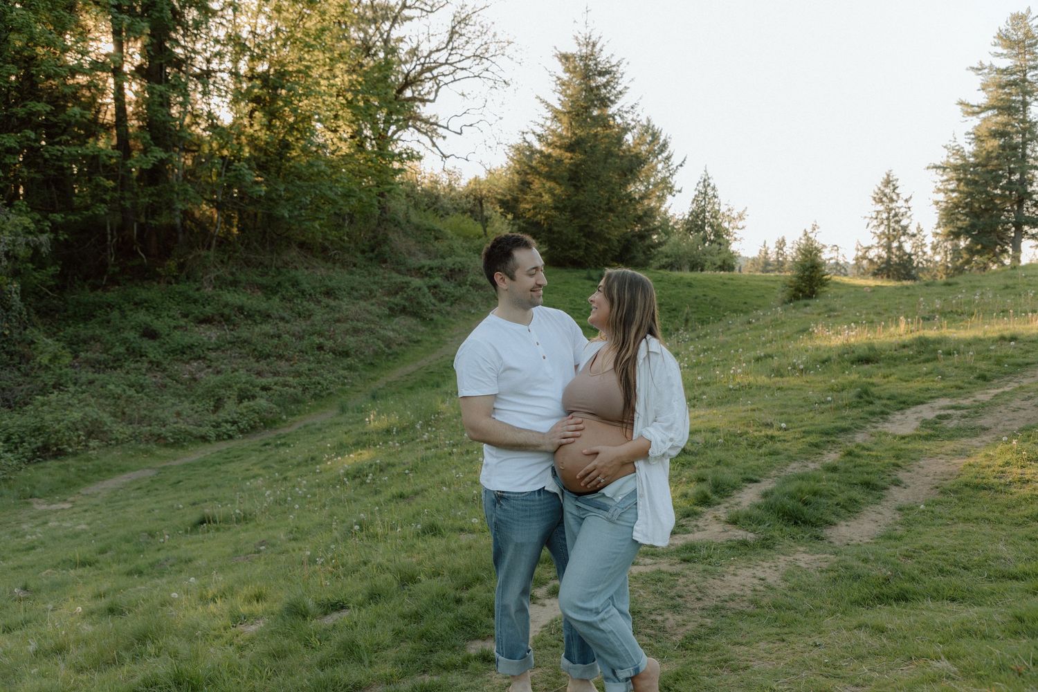 Expecting couple embraces while standing on grassy hillside at sunset.