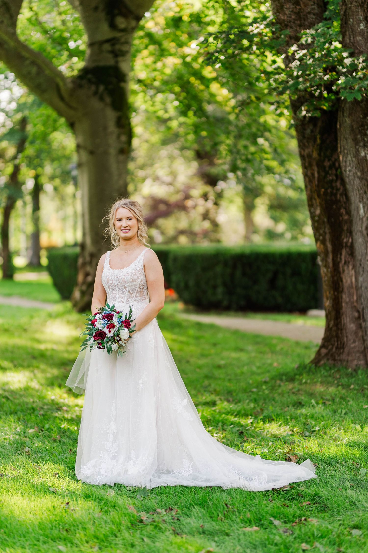 A bride in a white lace gown poses with purple bouquet in a garden setting.
