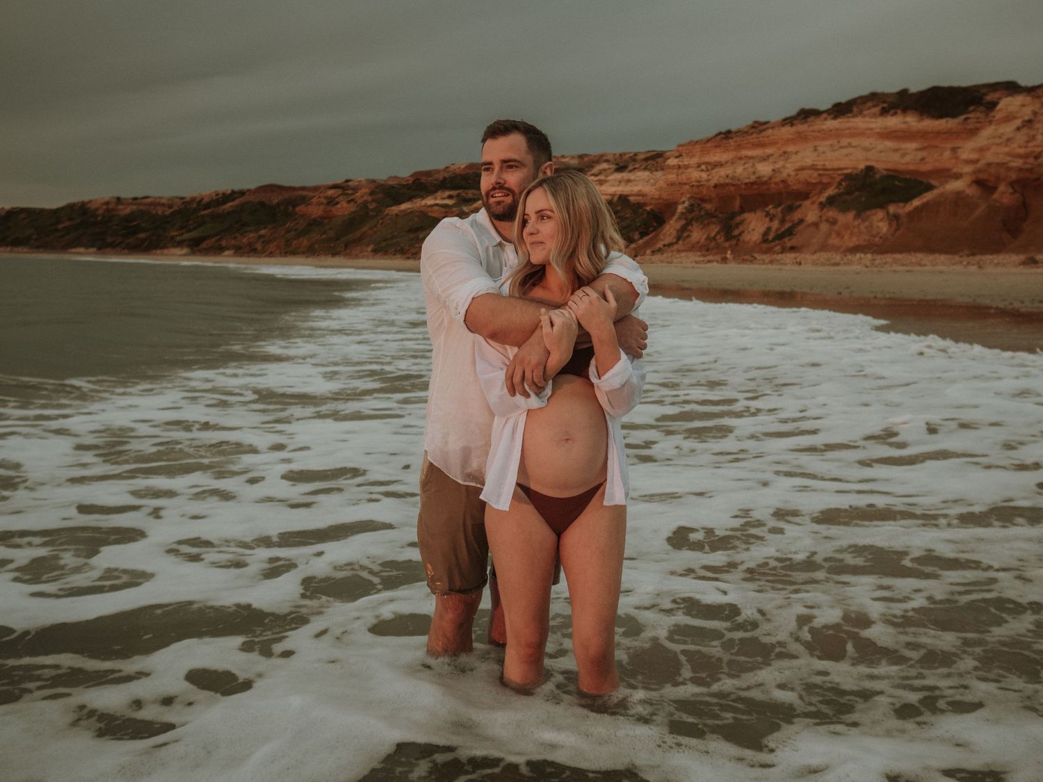 A joyful couple plays in ocean waves with red cliff coastline in the background.
