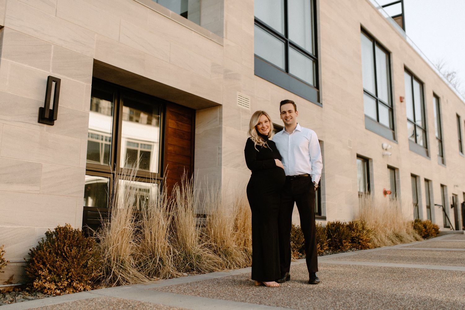 A couple poses together outside a modern building with glass windows and ornamental grasses.