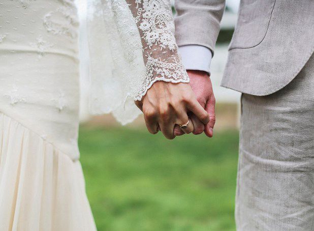 A couple wearing wedding attire holds hands in a close-up shot with green grass in the background.