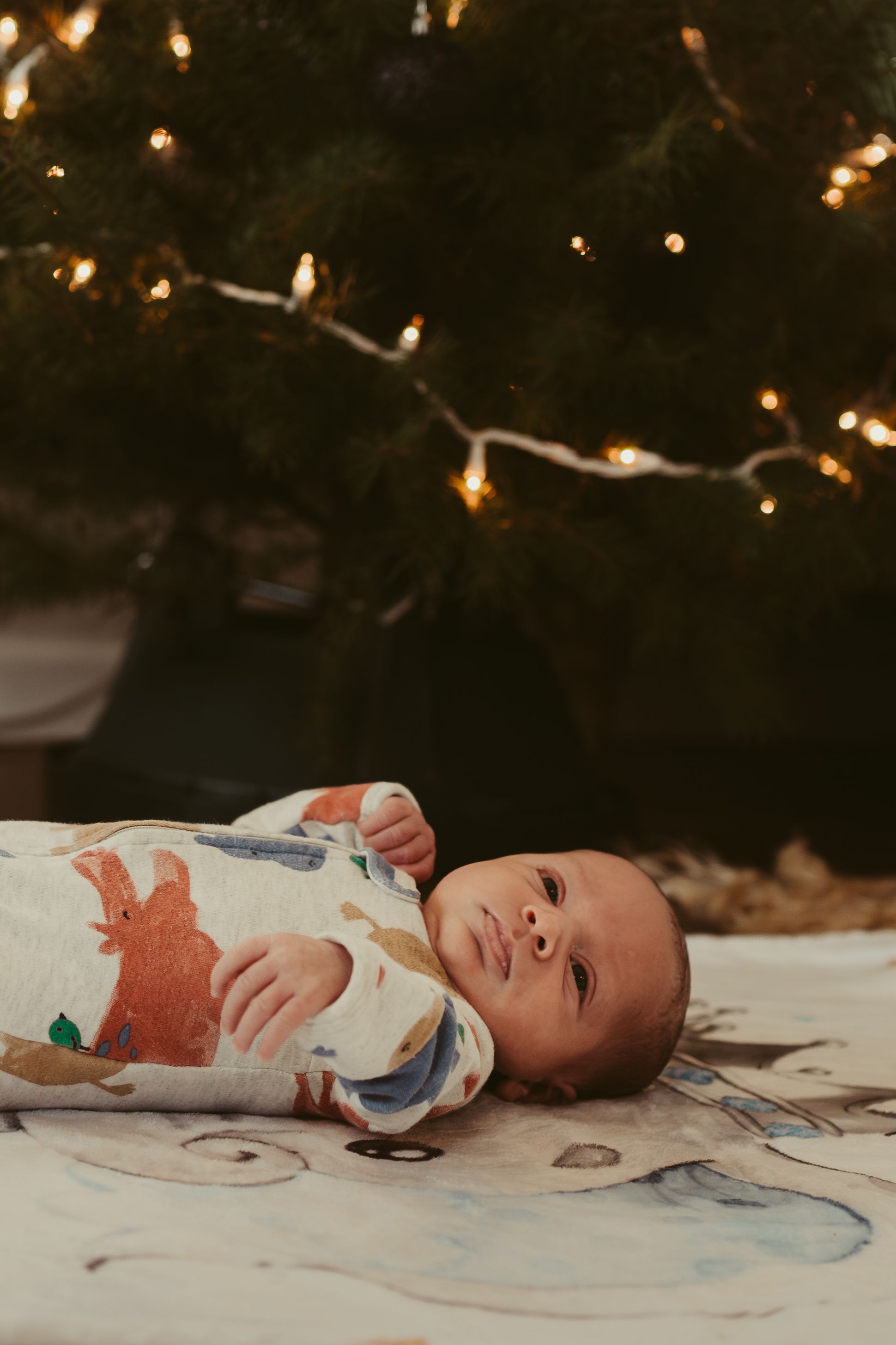 A newborn baby in a patterned onesie lies under twinkling Christmas lights.
